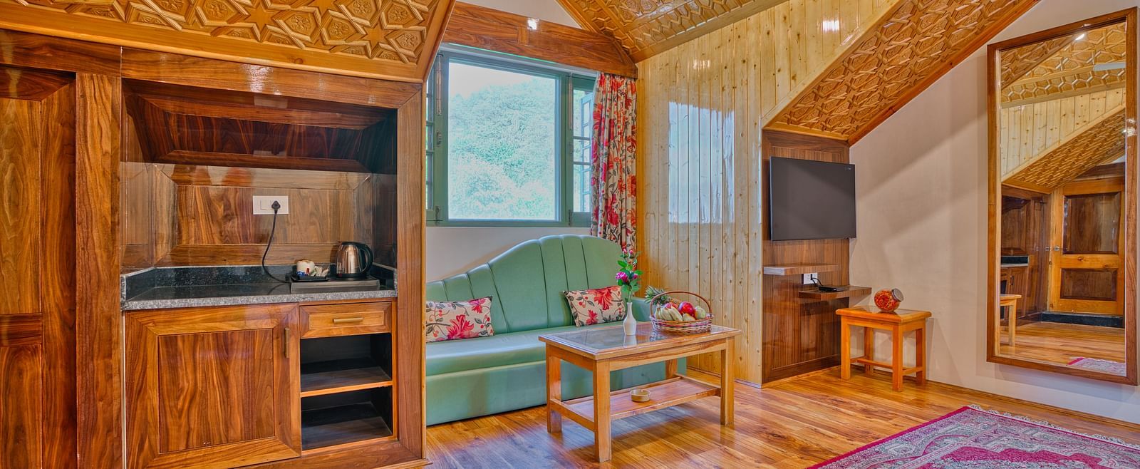 Interior of a room at Royal Comfort Regency showing a wooden kitchenette, seating area, sloped ceiling and large window with natural light.