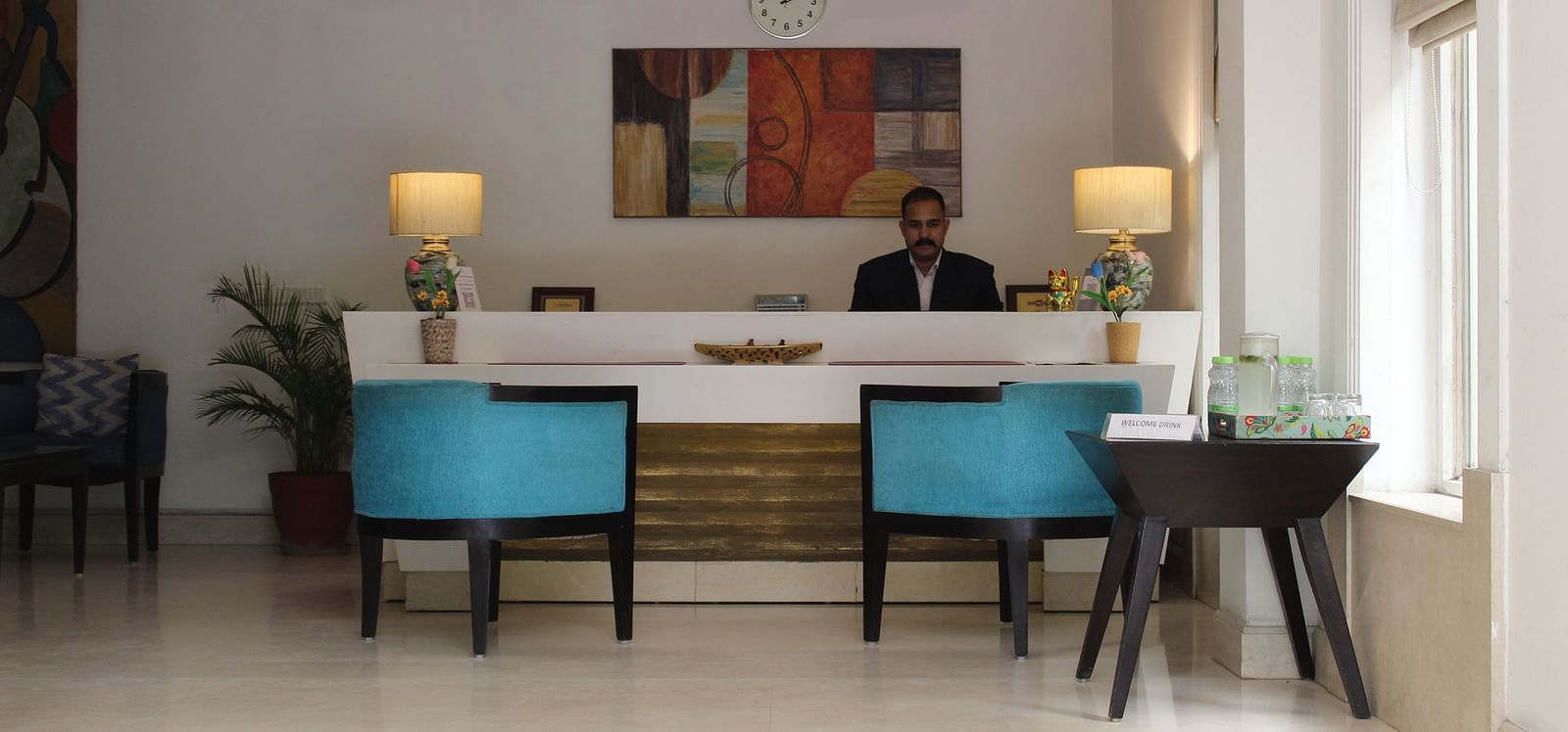 An image of a reception lobby featuring a reception desk where a reception is standing and in front of them there are two chairs placed along with a table at the corner of the room - Inde Hotels Vista Woods