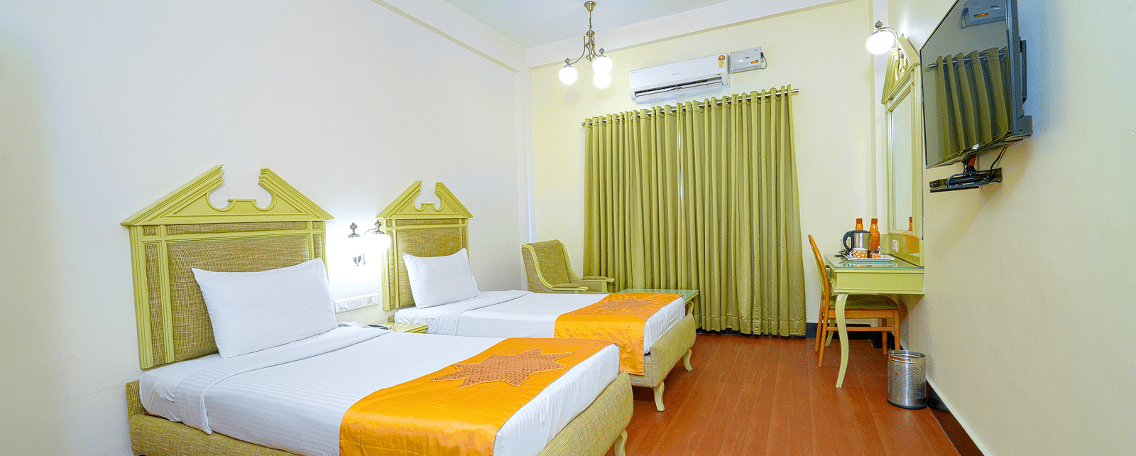 A twin hotel room with light wooden flooring, green headboard, and mustard accents, offering a cozy atmosphere at Greens Inn - Cozy Twin Rooms in Ramoji Film City.