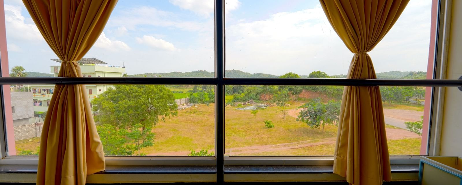 A scenic view from a hotel room window with yellow drapes overlooking a green landscape at Greens Inn - Scenic Views in Ramoji Film City.