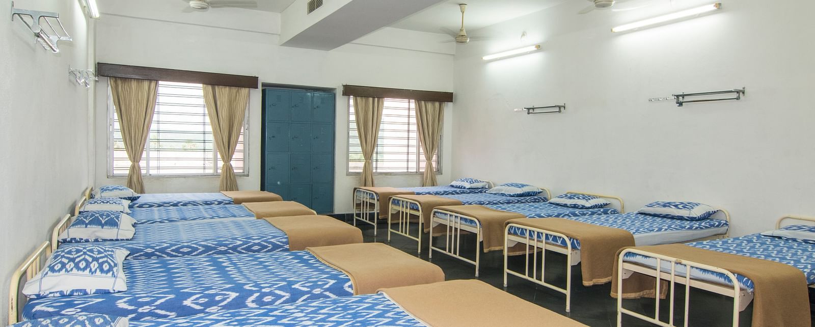 Beds with bedding are arranged in two rows in a room with two windows at Hotel Sahara - Comfortable Dormitory in Ramoji Film City, Hyderabad.
