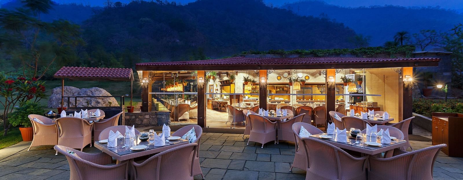 An outdoor restaurant with a view of mountains at dusk. Tables and chairs are set up, a covered area with warm lighting in the background.