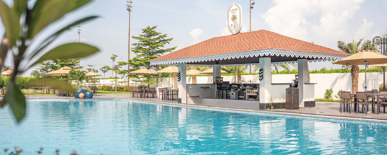 Sunny daytime view of the swimming pool with a red-roofed bar or pavilion and lush greenery in the foreground at Hotel Hukam's Lalit Mahal.