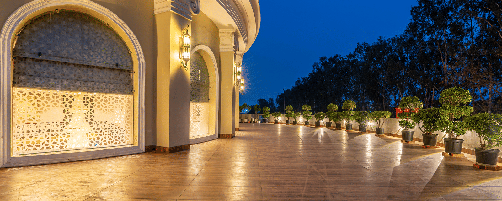Curved, illuminated walkway at night with decorative wall panels, a polished floor, and potted plants lining the path at Hotel Hukam's Lalit Mahal.