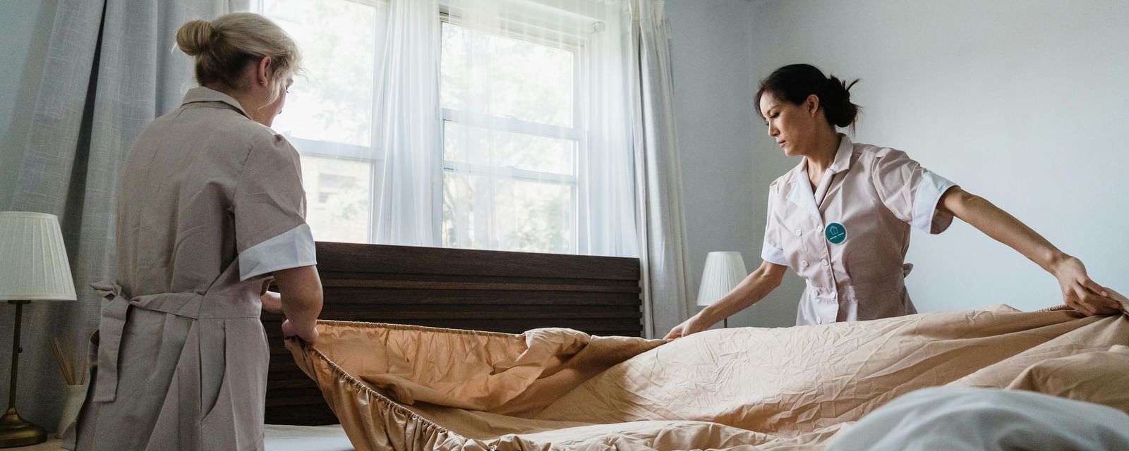 2 women making a bed in a guest room, with windows, curtains and bright atmosphere.