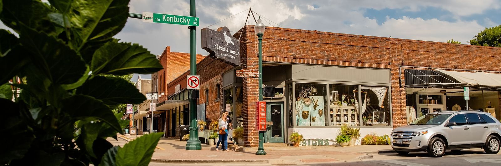 A street lined with shops, trees, a traffic light, and cars parked along the road, viewed from a landscaped corner.- The Grand Hotel & Ballroom, McKinney