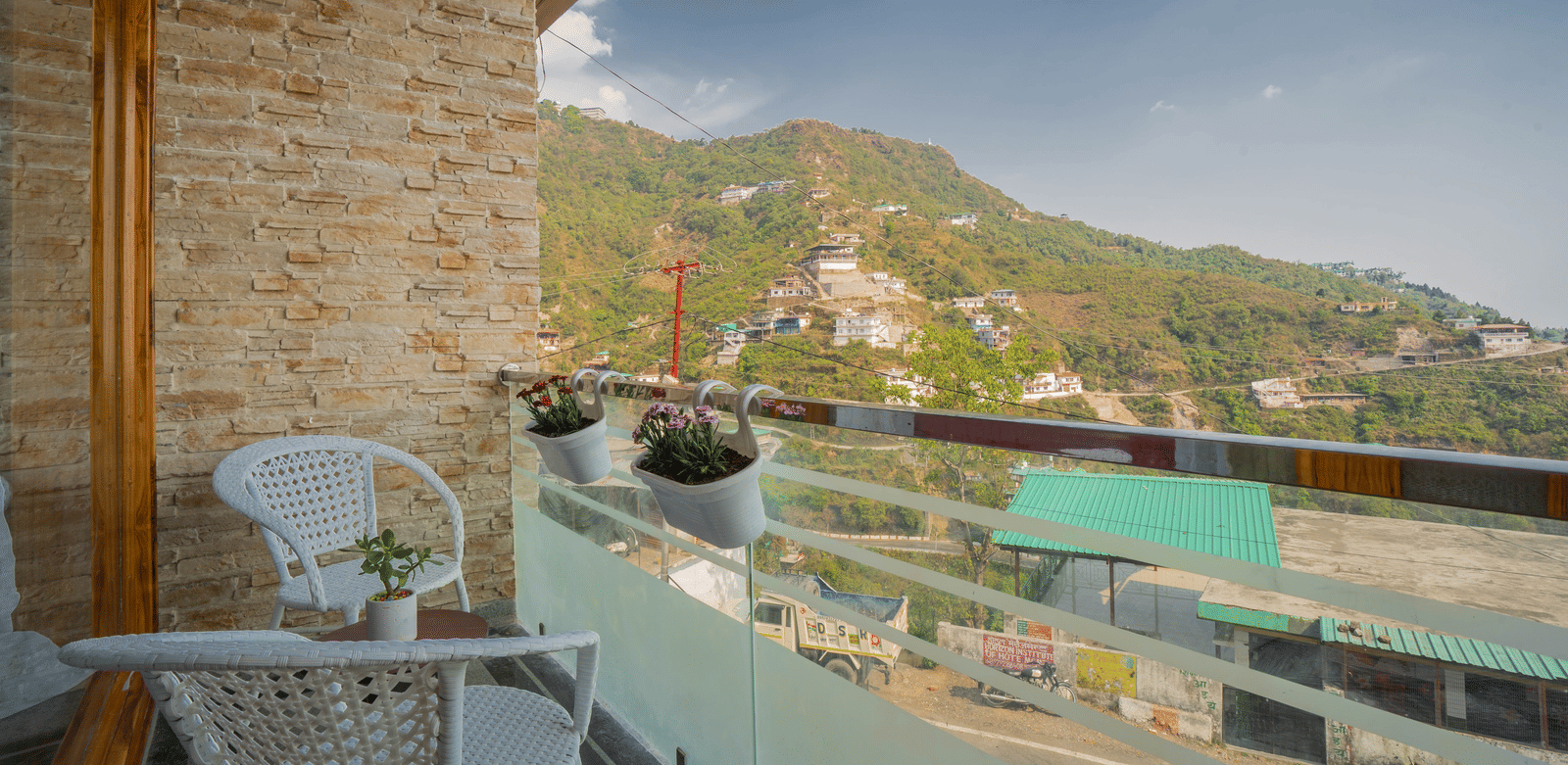 A balcony at Perfectstayz Value Paradise, Mussoorie, featuring a white chair, small table, and potted plants overlooking scenic hillside views and rooftops.