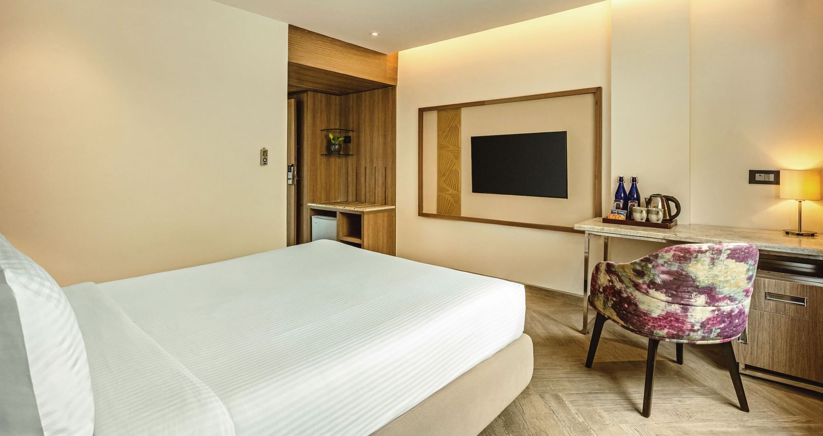 A wide-angle view of a room with a bed and a television at Mango Hill Central, Chennai. A desk with a chair is in the foreground.