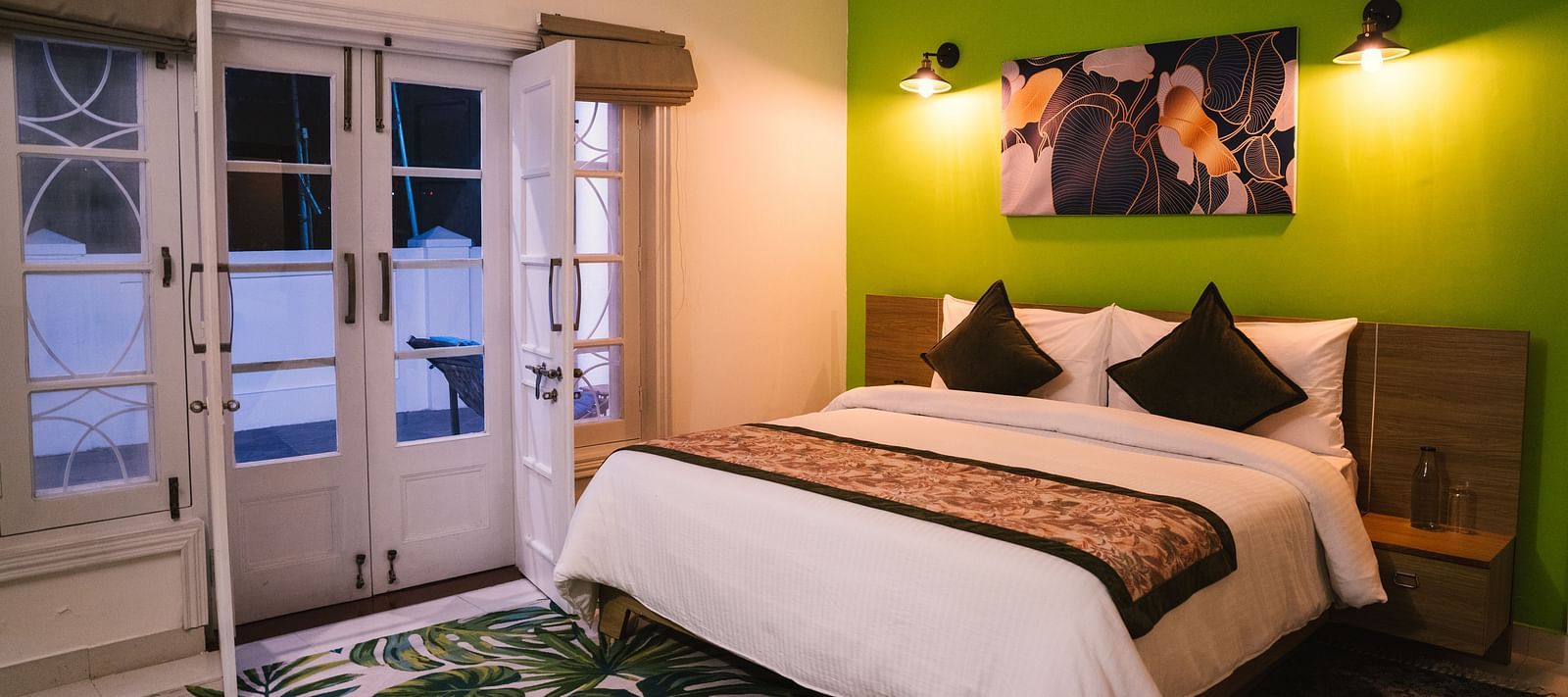 A spacious room at BluSalzz Villas - The Green Roof, Bhimtal, featuring a large bed with a photo frame above the headboard, night lamps glowing on either side, a carpet, and a door leading to the balcony.