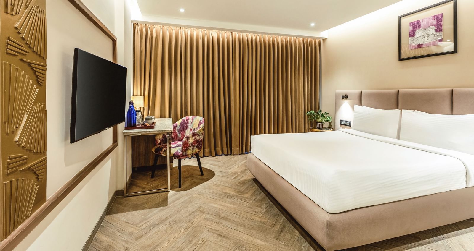 A wide-angle view of a room with a large bed and a television at Mango Hill Central, Chennai. A desk and chair are visible on the right.