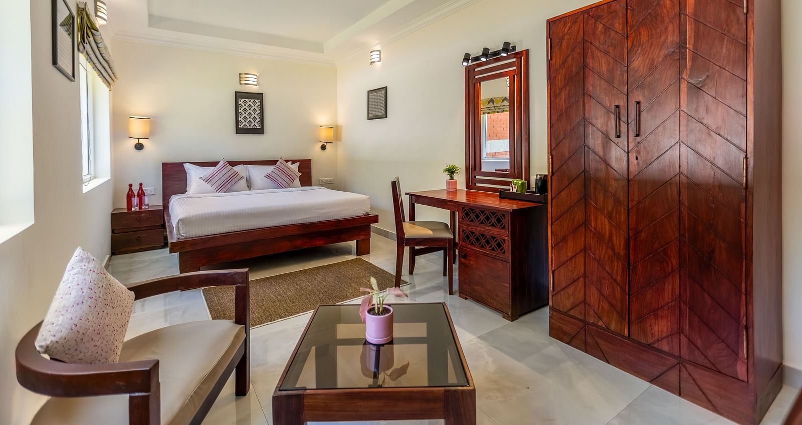 A bright, clean hotel room with a bed, a wooden desk and chair, a large wooden wardrobe, and a seating area with a glass-topped coffee table - Mango Hill Resort, Coimbatore