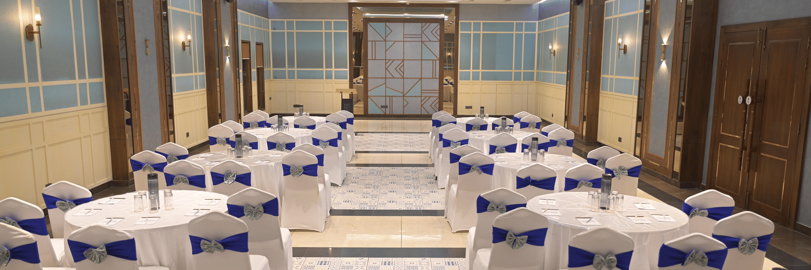 A straight, centred view of the banquet hall with round tables and blue-accented chairs at Hotel Sonar Bangla Mayapur.