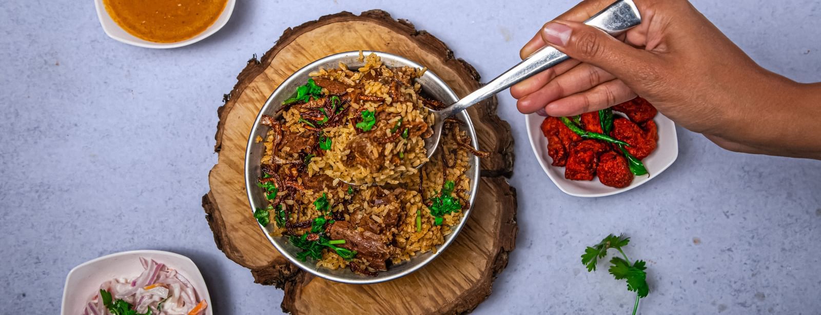 A hand scooping from a bowl of richly garnished Indian curry served on a wooden platter, accompanied by bowls of chutney placed on a light blue surface at The Hamuse Hotel, Kodaikanal.