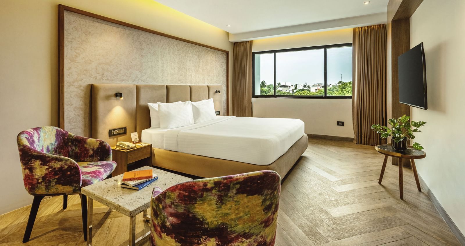 A wide-angle view of a room with a large bed at Mango Hill Central, Chennai. There are a chair and a small table in the foreground.