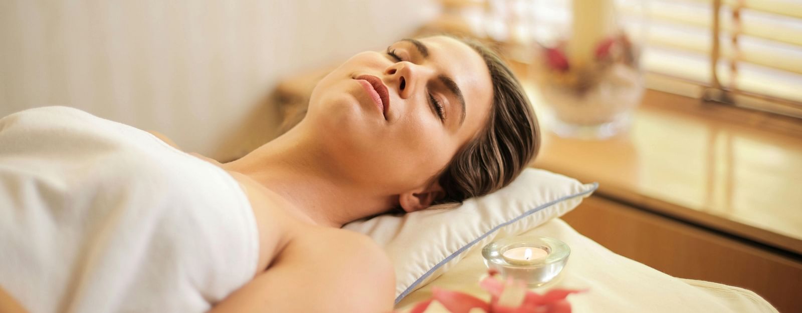 A woman lay on a spa bed, wrapped in a white towel with candles and flowers visible nearby.