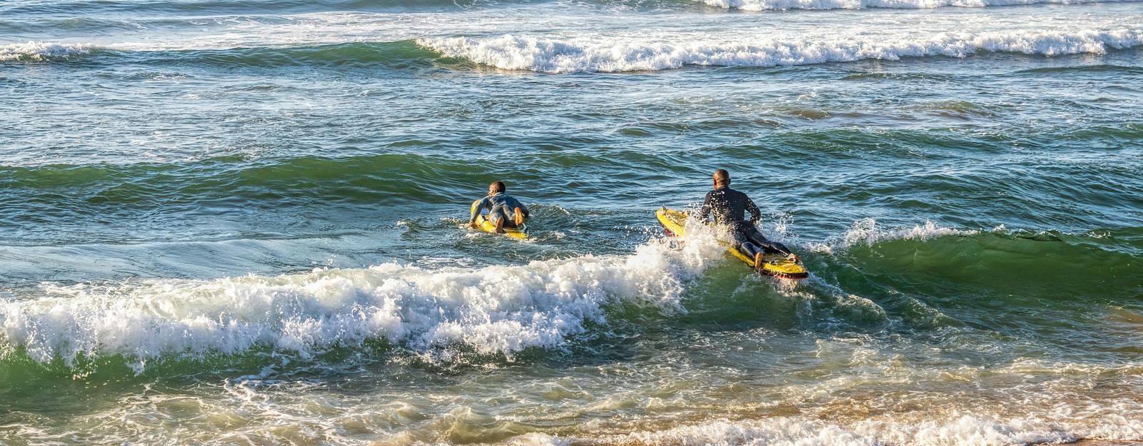 Two people surfing on breaking waves near a sandy beach.