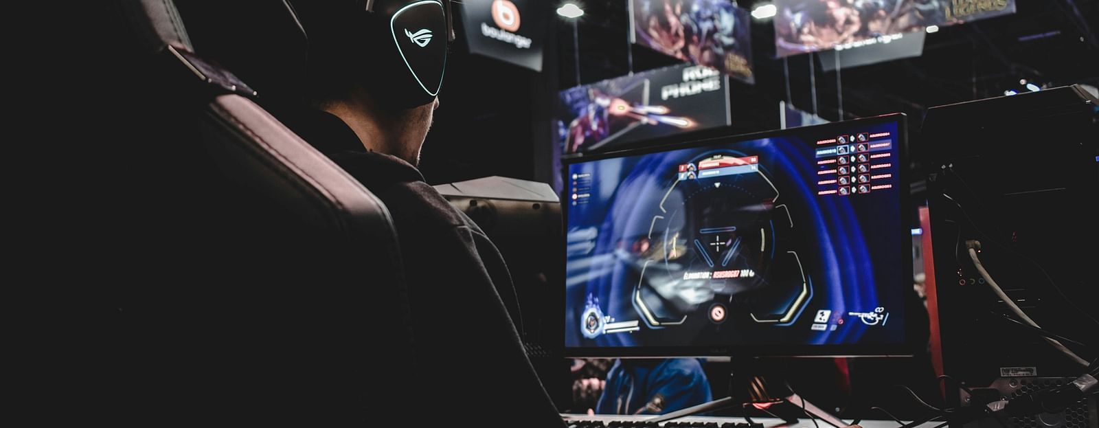 Side view from the back of a man wearing gaming equipment and playing games on a machine. There are screens in the background.