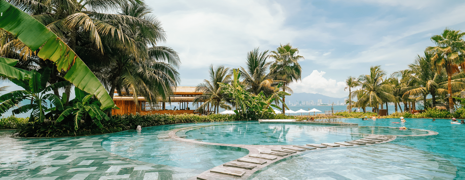 Swimming pool surrounded by palm trees at Alibu Resort in Vietnam, featuring several layers and narrow pathways in it where people can walk and go to different sections of the pool.