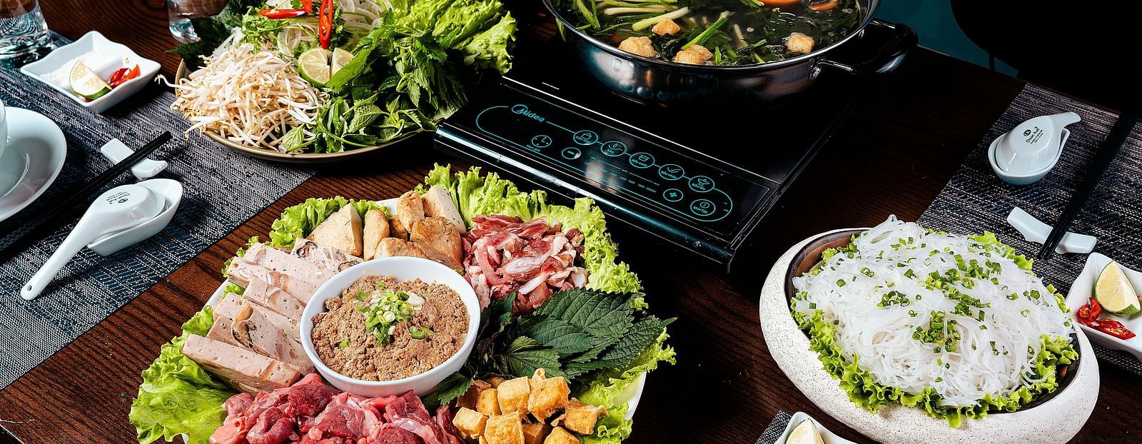 A table set with a variety of dishes including sliced meats, vegetables, noodles, and a pot of broth, ready for a meal.