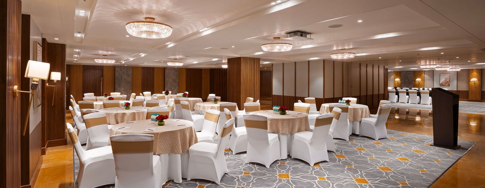 A spacious banquet hall with round tables covered in tablecloths and chairs. A podium in the background, and the room is decorated with chandeliers and wall lamps.