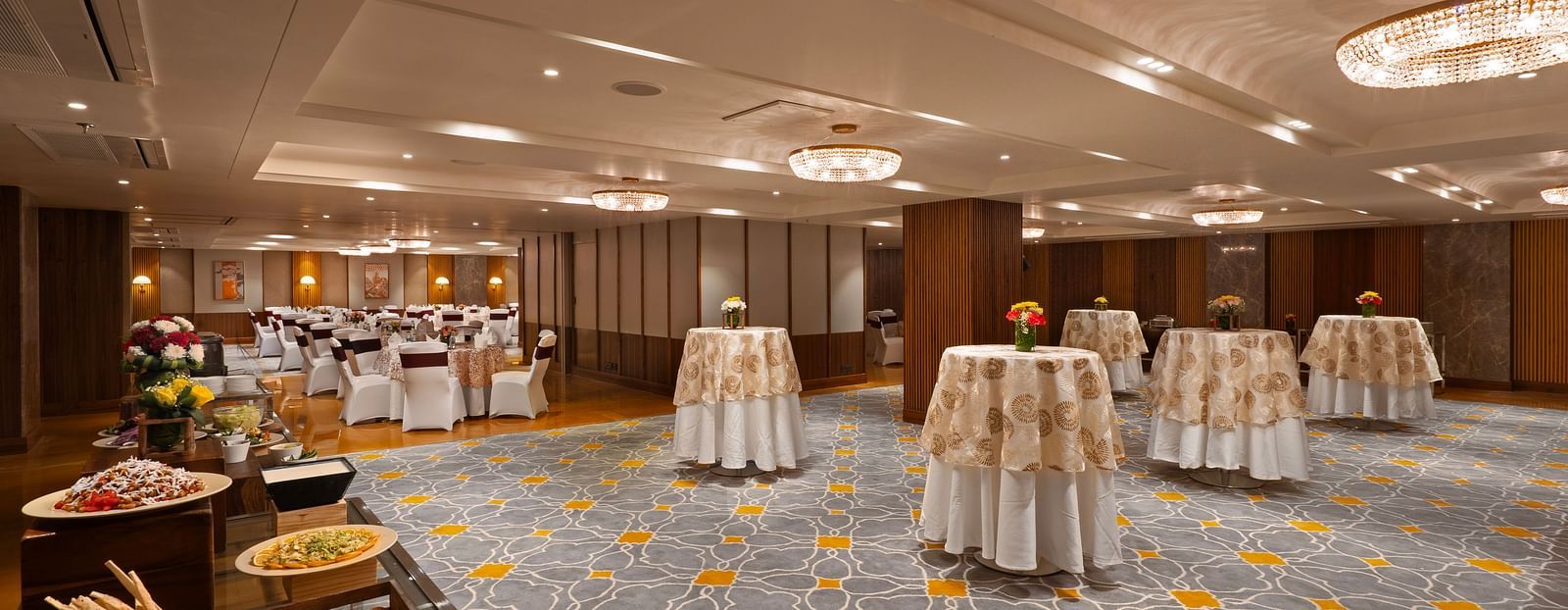 This image depicts a spacious and elegant banquet hall, The room features high ceilings, ornate chandeliers, and a plush carpeted floor with several round tables are arranged throughout the space.