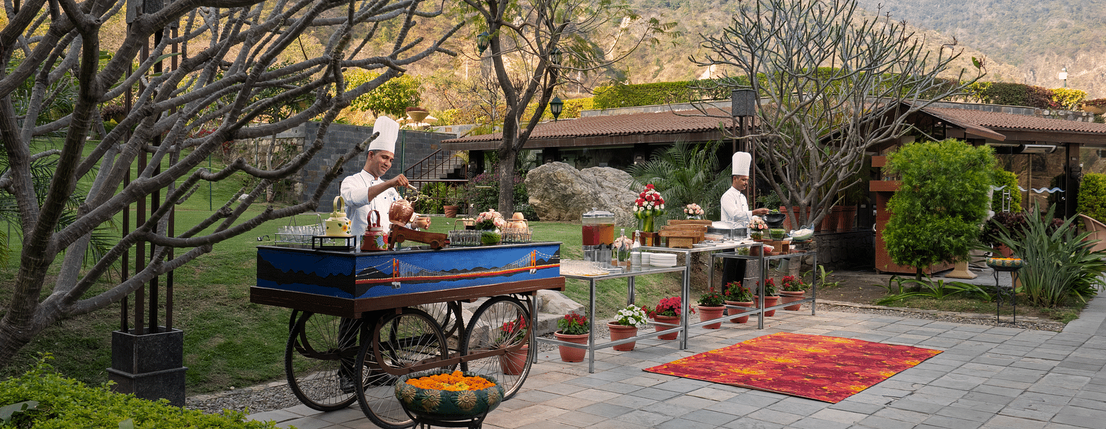 Two chefs are preparing food at outdoor food stations. There are mountains in the background and lush surrounding the scene.