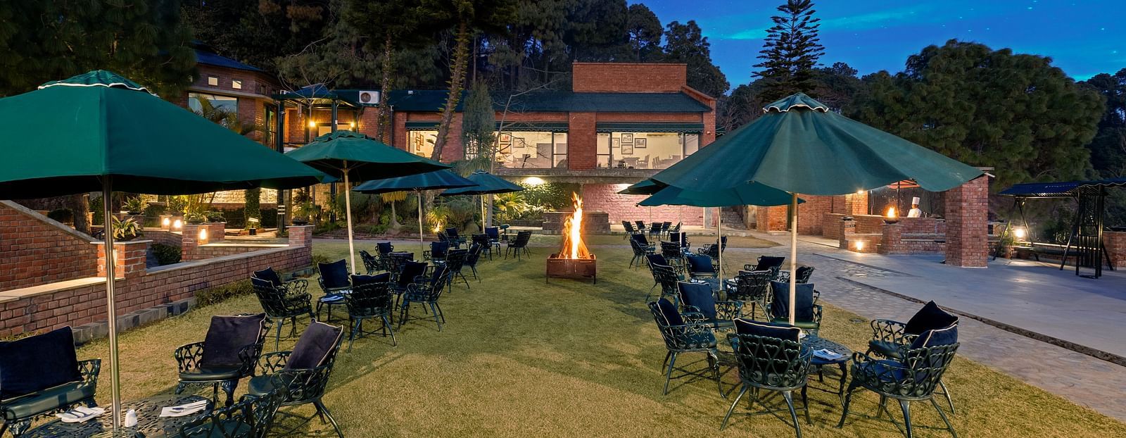 An outdoor lawn with an umbrella-shaped roofs providing shade, surrounded by chairs arranged for seating -  Baikunth resort, kasauli 