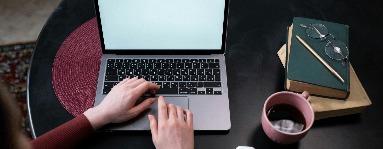 A person is using a laptop placed on a black table. To the right of the laptop, there’s a coffee mug and next to it are spectacles and a diary.