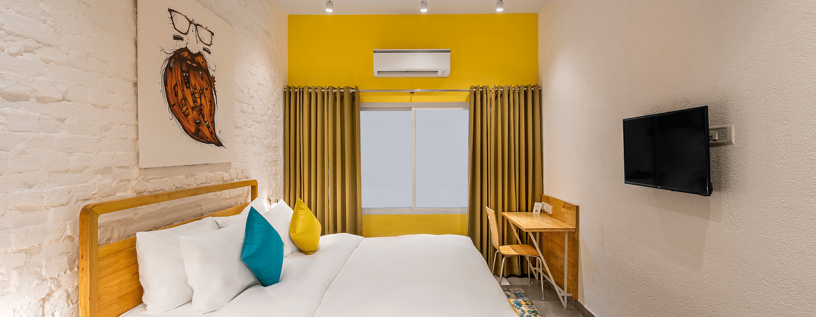 An side angle interior image of the standard room at Bedzzz Varanasi showcases a spacious layout, featuring a comfortable double bed, a cozy floor mat, a stylish study table and chair, a charming coffee table, elegant curtains, an air conditioner for a ref
