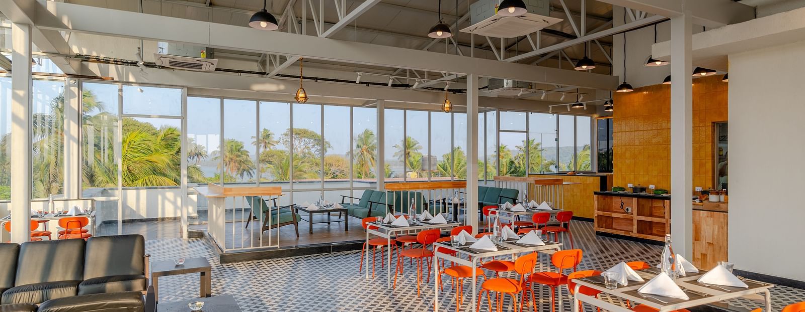  spacious and airy restaurant with industrial-style exposed ceiling, large windows letting in natural light, and a color palette of orange and grey, providing a relaxed dining atmosphere  at Bedzzz Xclusiv Morjim, Goa
