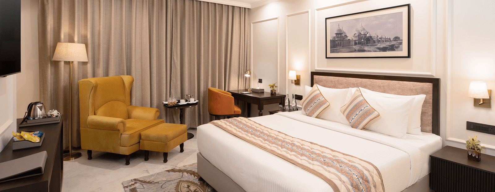 An image of a bedroom with bedside table, photo frame hanged on the wall, work desk with sofa chair, and a flat TV screen - Brij Eternity, Vrindavan