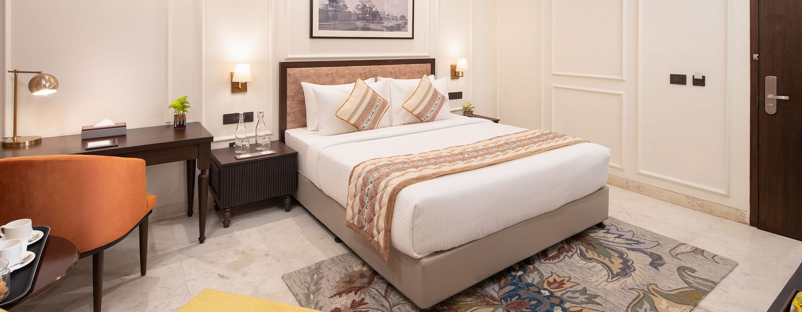 An image of a bedroom with bedside table, photo frame hanged on the wall, work desk with sofa chair, and a flat TV screen - Brij Eternity, Vrindavan