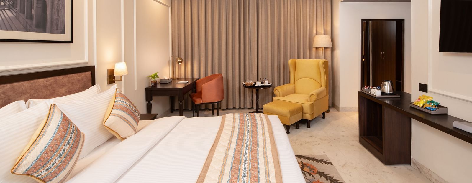 An image of a bedroom with bedside table, photo frame hanged on the wall, work desk with sofa chair, and a flat TV screen - Brij Eternity, Vrindavan