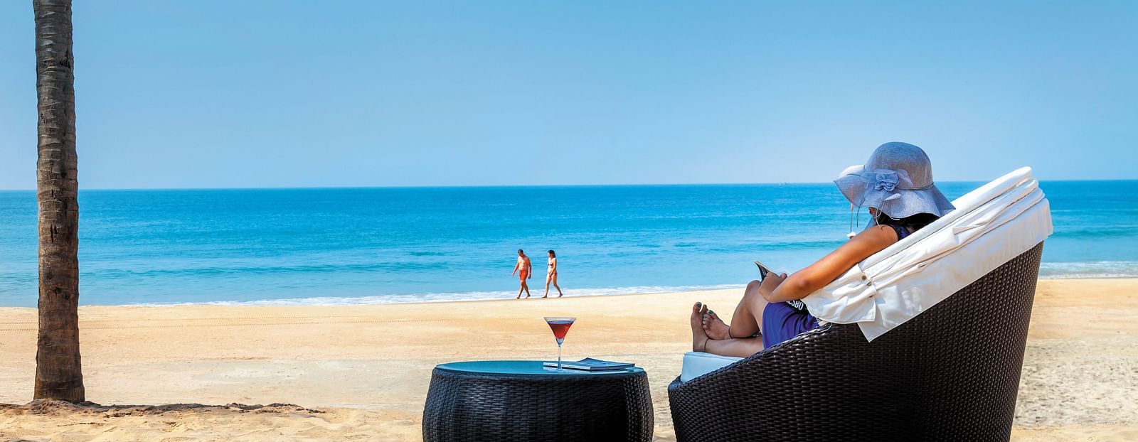 a woman sitting on the chair with a drink on the table on the beach - Caravela Beach Resort Goa.