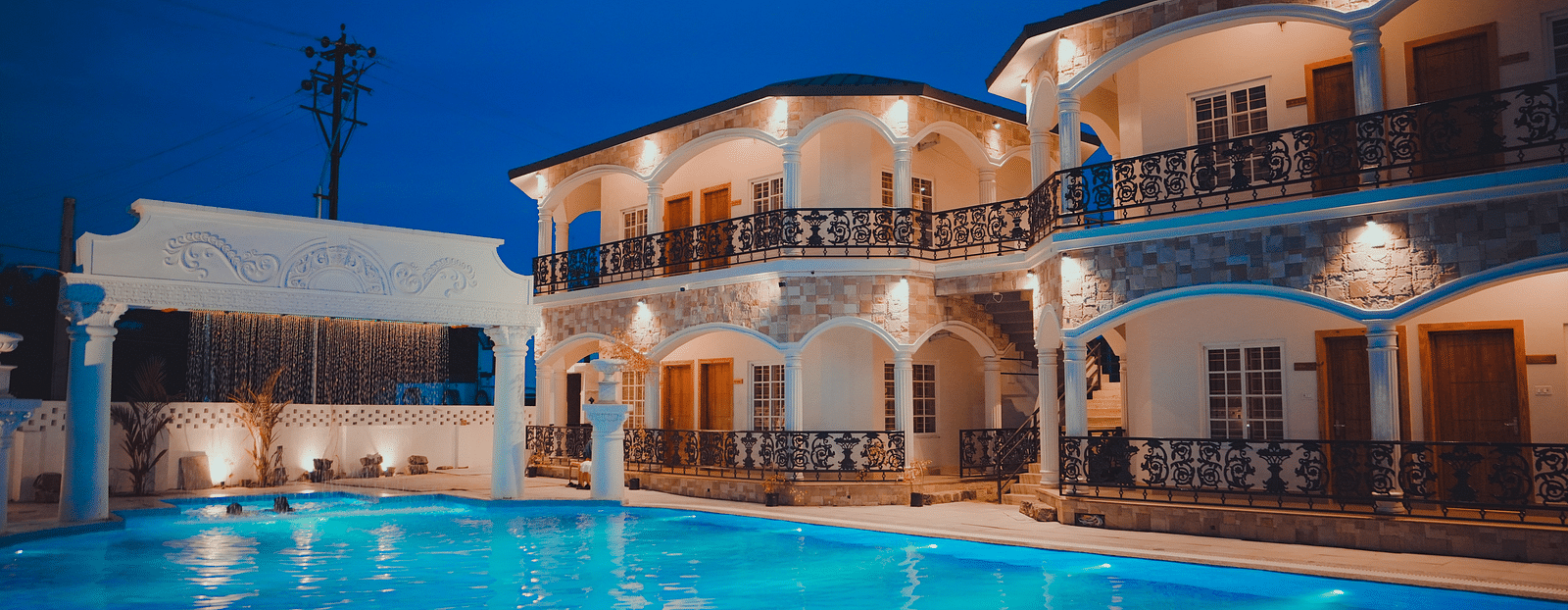 facade image of dip holidays hotel in birbhum with exterior lights switched on with an expansive blue swimming pool in the foreground
