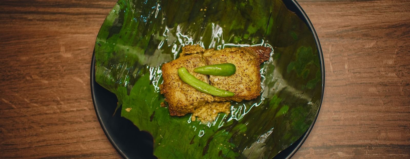 image of a fish preparation made with mustard steamed inside a banana leaf topped with two green chillies - Dip Holidays, Birbhum