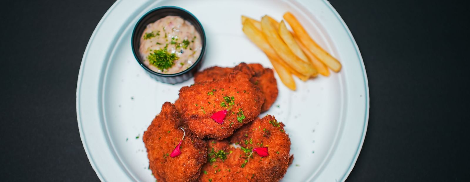 A white plate served with three pieces of crispy fried chicken, a side of golden French fries, and a small bowl of creamy dip, photographed on a dark background at D Café in Dip Holidays.