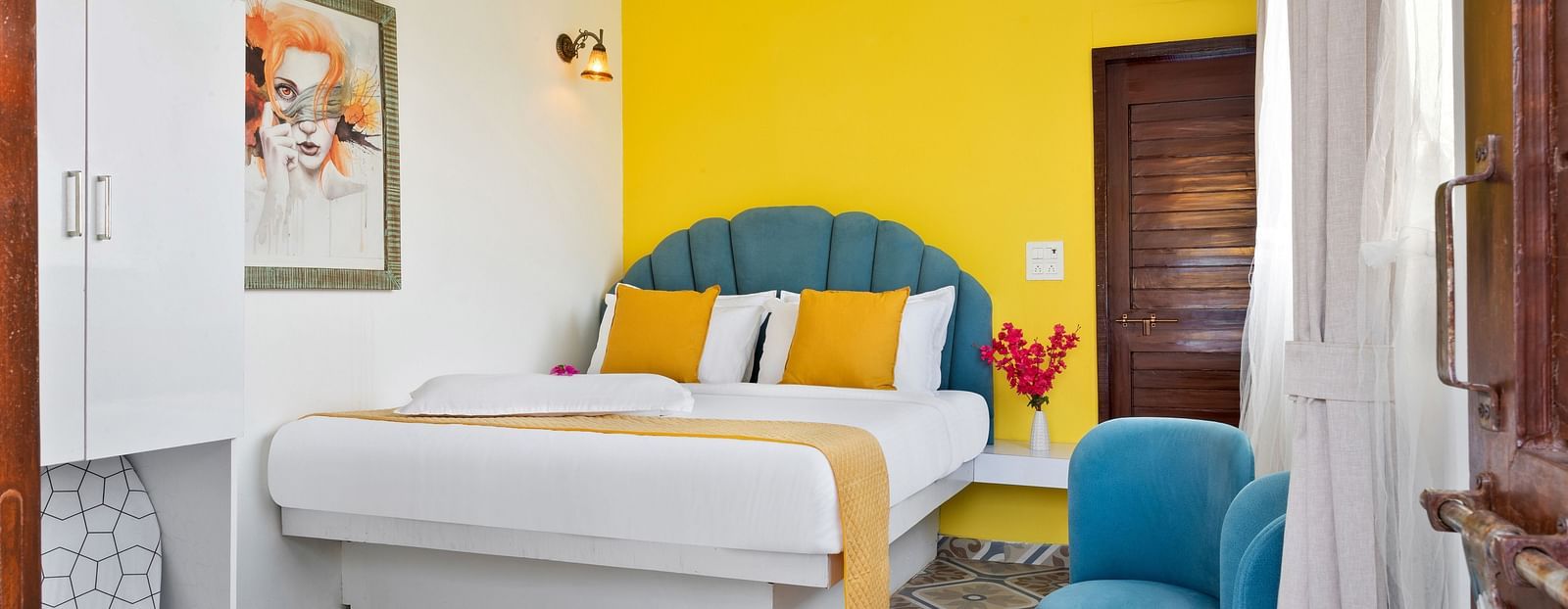 A cosy bedroom with a accent wall, a bed with white and mustard linens, and teal armchairs next to a small table.