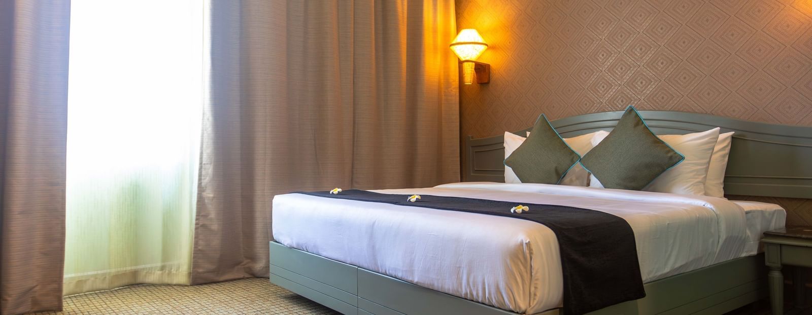 A hotel room featuring a bed with white and green bedding, a bedside lamp, and a window with curtains | Shilton Hotels