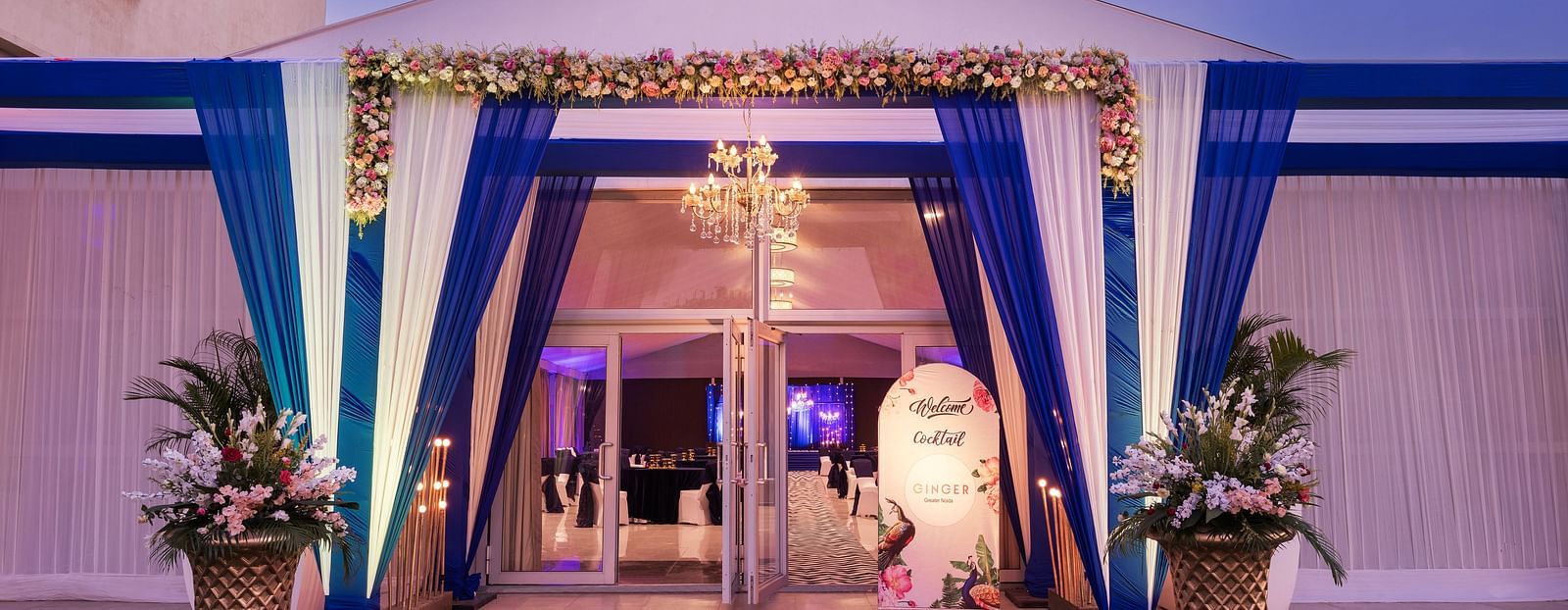 an entrance to a hall with a sign board beside the entry gate and blue and white curtains in front of the entrance - The Hideaway Banquets & Lawns, Greater Noida