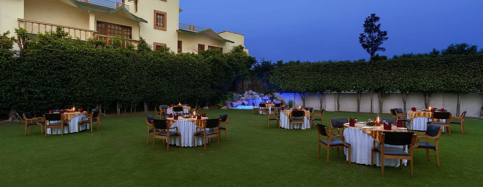An open lawn with tables and chairs arranged in a circle, each adorned with decorative settings and a small lamp, featuring a lit-up fountain in the corner