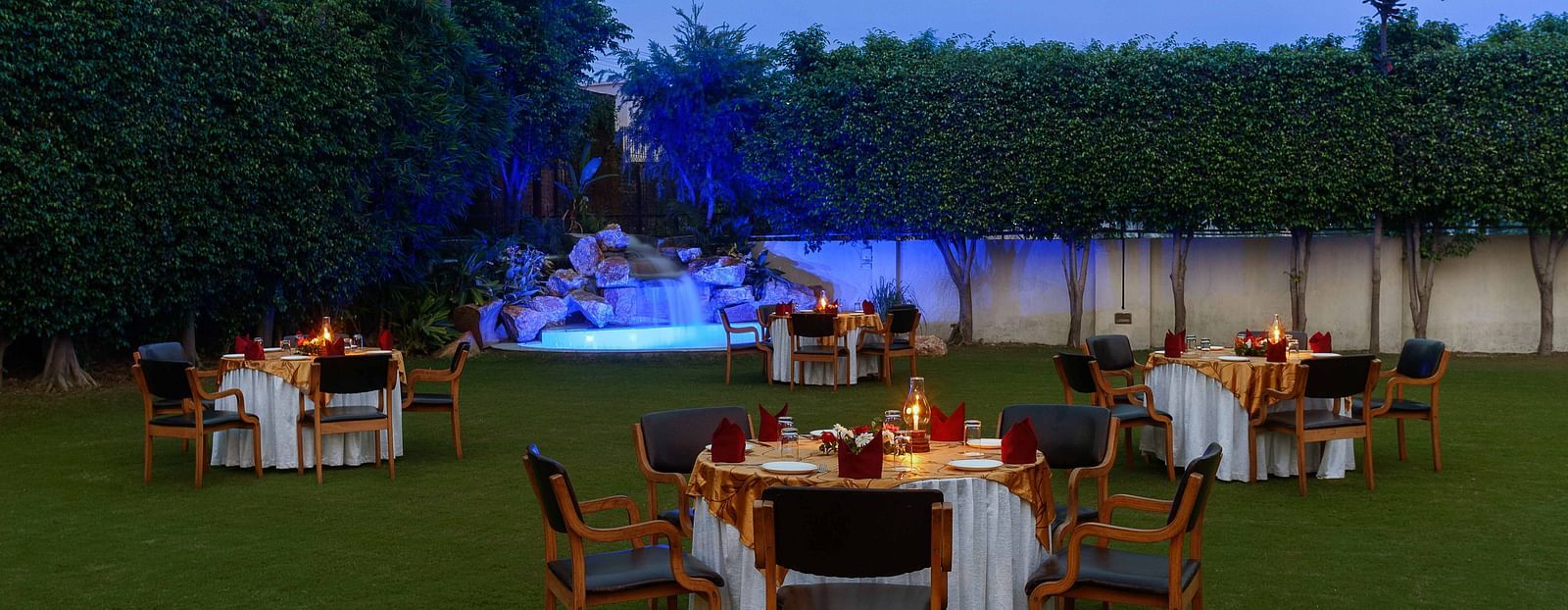 An open lawn with tables and chairs arranged in a circle, each adorned with decorative settings and a small lamp, featuring a lit-up fountain in the corner