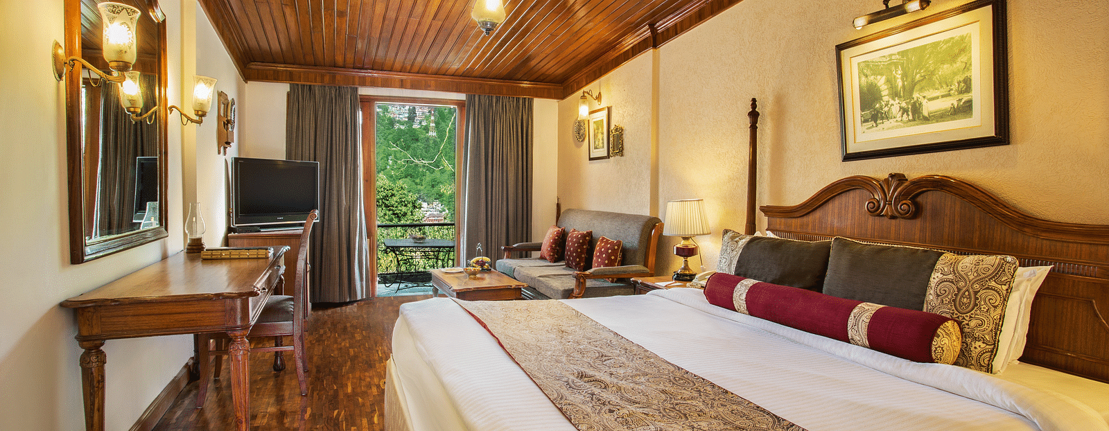 An image of a hill view room in naini retreat, nainital, with a king-size bed, wooden furniture, and a bay window overlooking a garden. The room features a ceiling fan, a television, and a small sitting area.