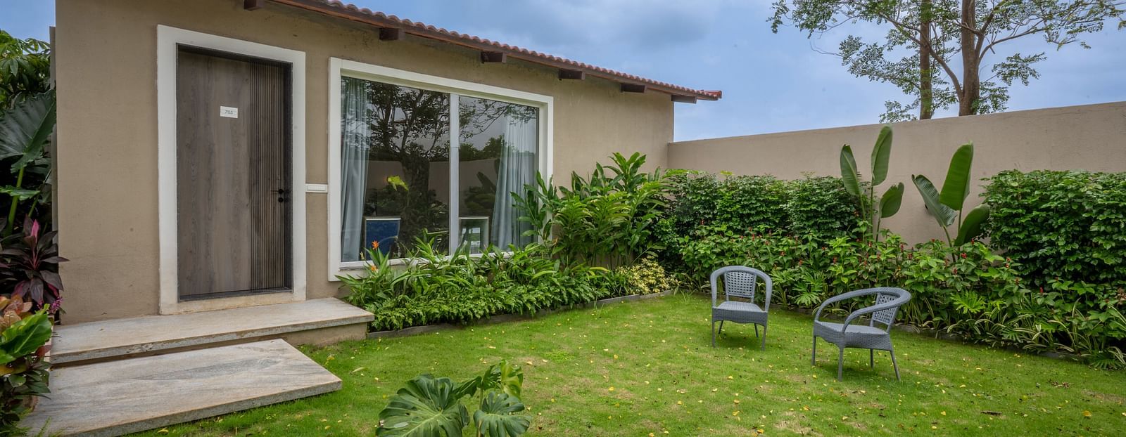 Ananta Garden Villa exterior showing the villa building, a lush green garden with outdoor seating, and cloudy sky.