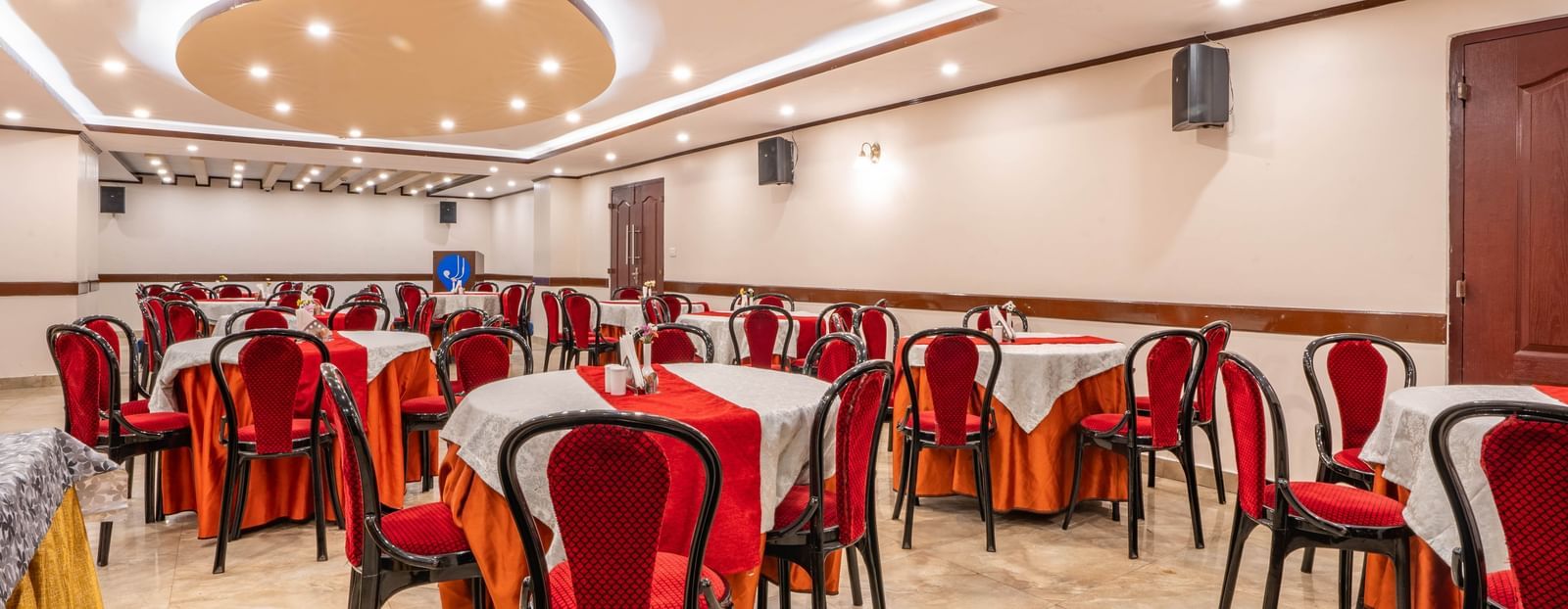 A spacious banquet hall at the Yashshree Jai Hotel & Banquet, featuring a setup with round tables, white cloths, and red chairs.