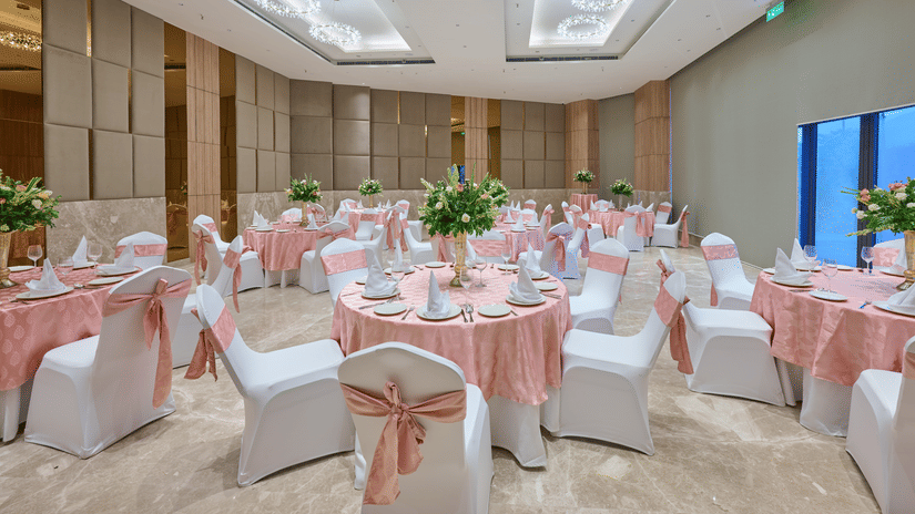 An event hall view at KPG Sarovar Portico, Raj Nagar Extension, Ghaziabad featuring round tables, chairs with bows, modern ceiling lights, and tile flooring.
