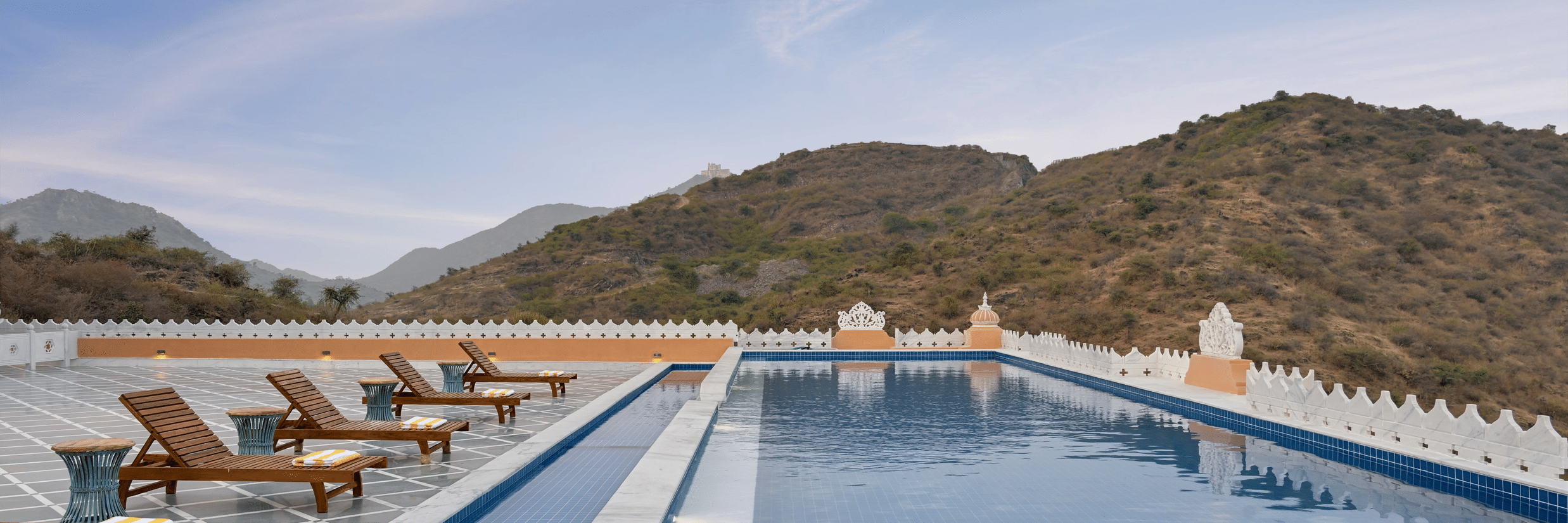 Swimming pool with loungers next to it and mountains in the background at Fateh Vilas Udaipur. Get access to the contact of Fateh Vilas by Fateh Collection for more information.