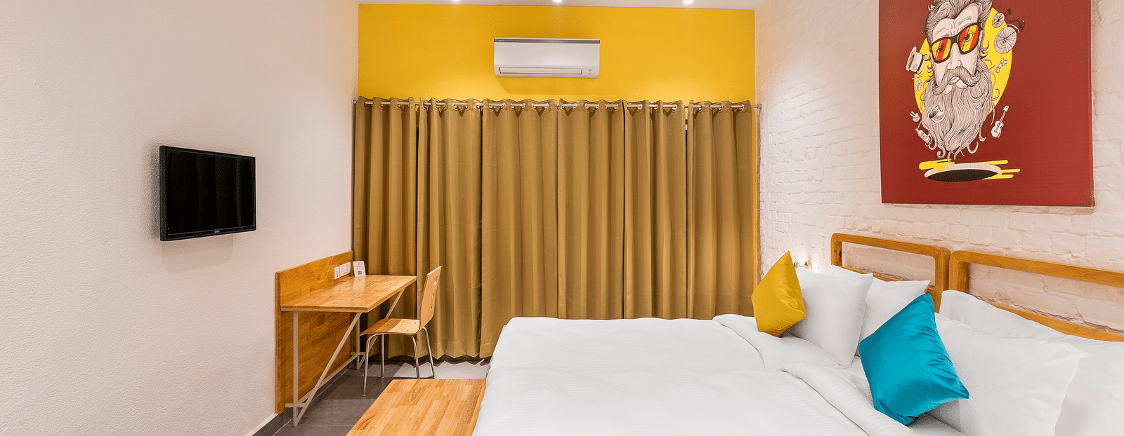 A side-angle interior image of the deluxe room at Bedzzz Varanasi showcases a spacious layout featuring a large bed, a window with elegant curtains, a TV, a decorative wall poster, ample lighting, and a cupboard for storage.