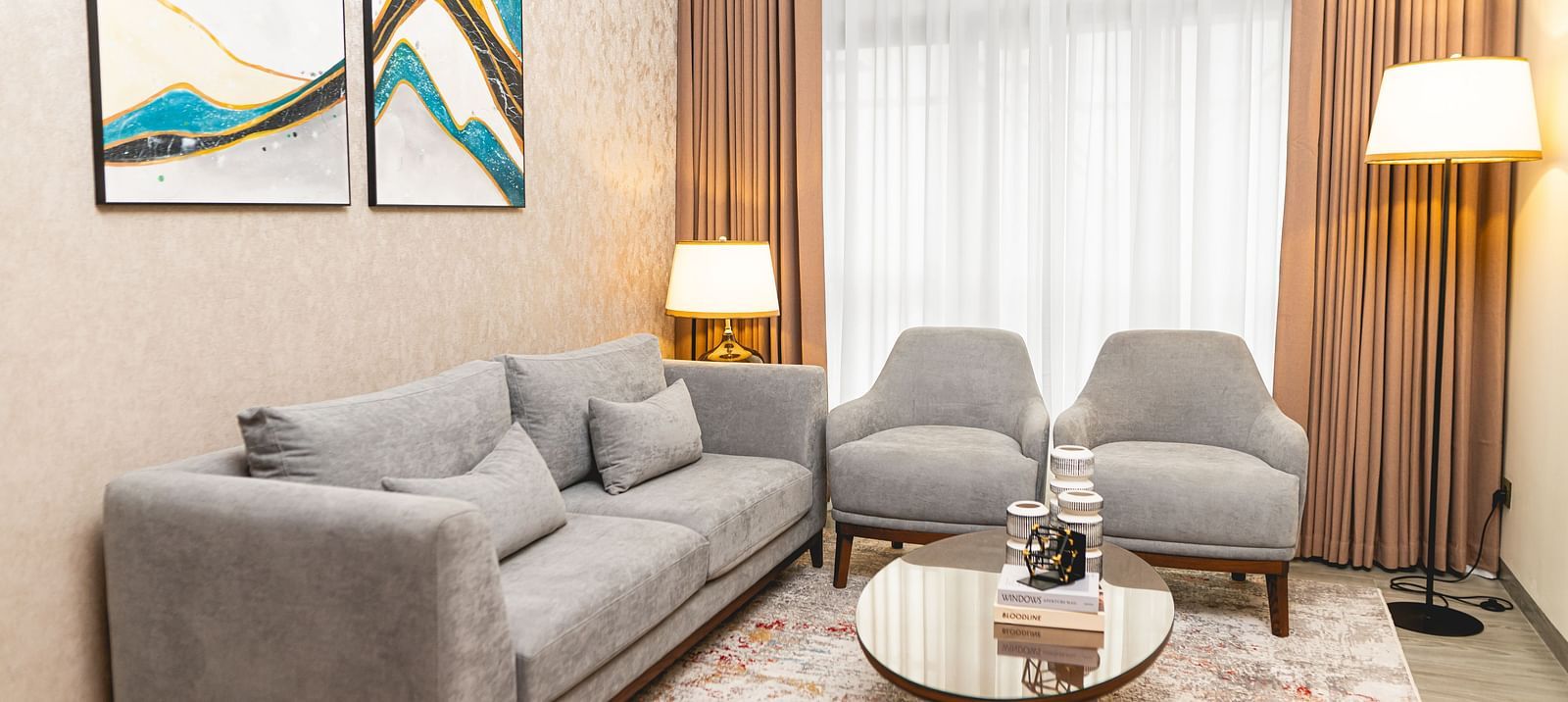 Room at KertelSuites with a sofa, two matching armchairs, a round coffee table, two floor lamps, a patterned rug, and abstract art on the wall. Large windows with sheer curtains allow natural light in.