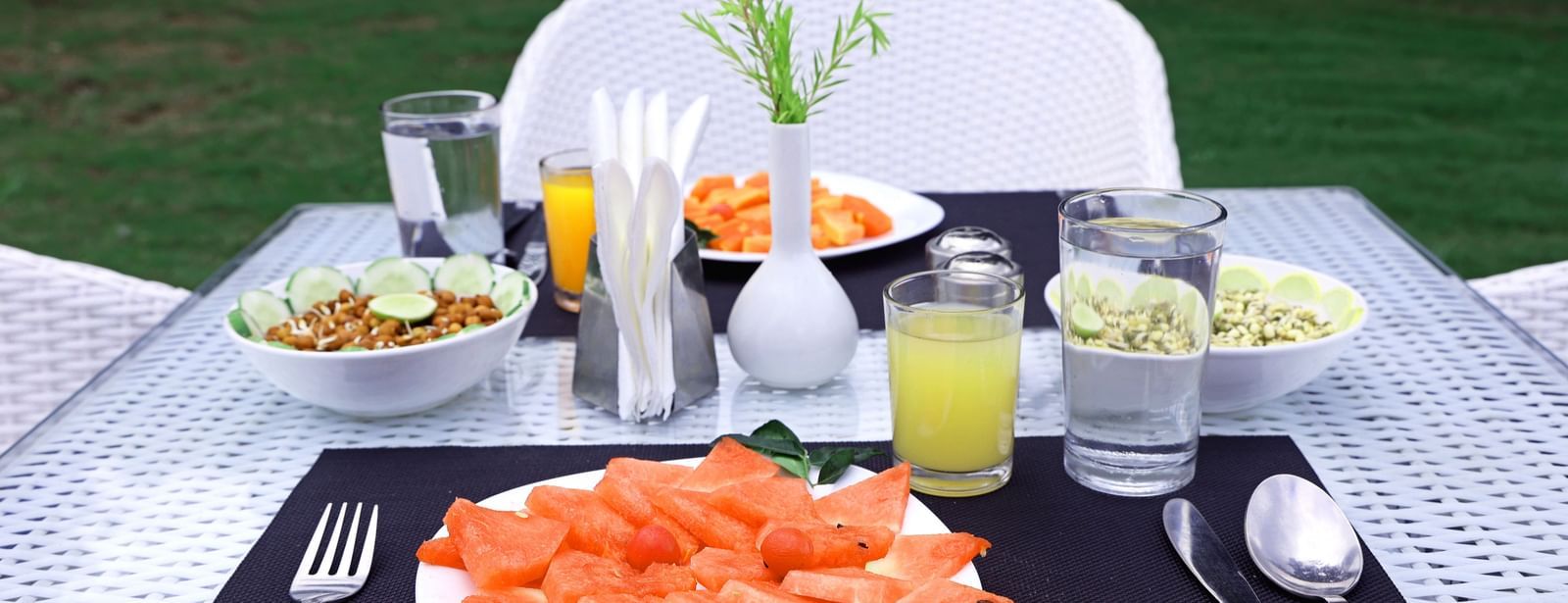 A neatly arranged outdoor dining table is set with fresh fruit, beverages, and cutlery at Corbett Nirvana Resort.